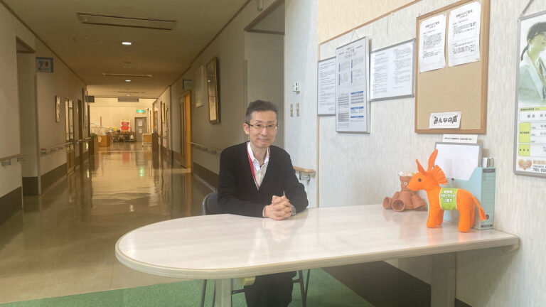 介護施設のロビー・受付エリア。スタッフが受付デスクに座り、掲示板や廊下が見える。